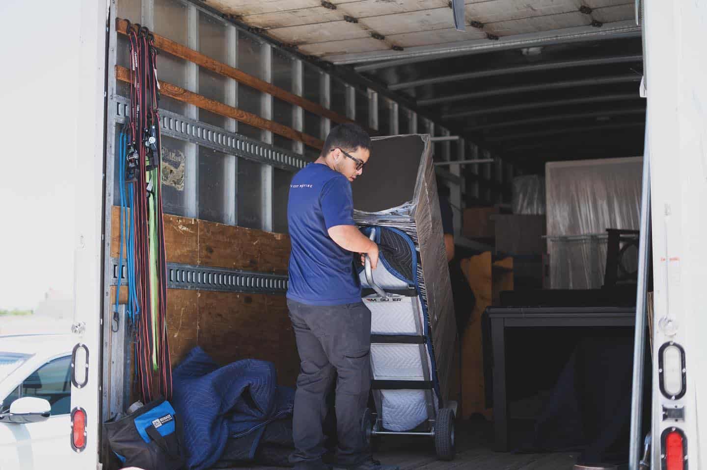 https://www.jp-moving.com/wp-content/uploads/2024/03/a-mover-in-a-blue-jp-moving-shirt-pushes-a-plastic-wrapped-item-on-a-dolly-inside-a-moving-truck-filled-with-furniture11.jpg